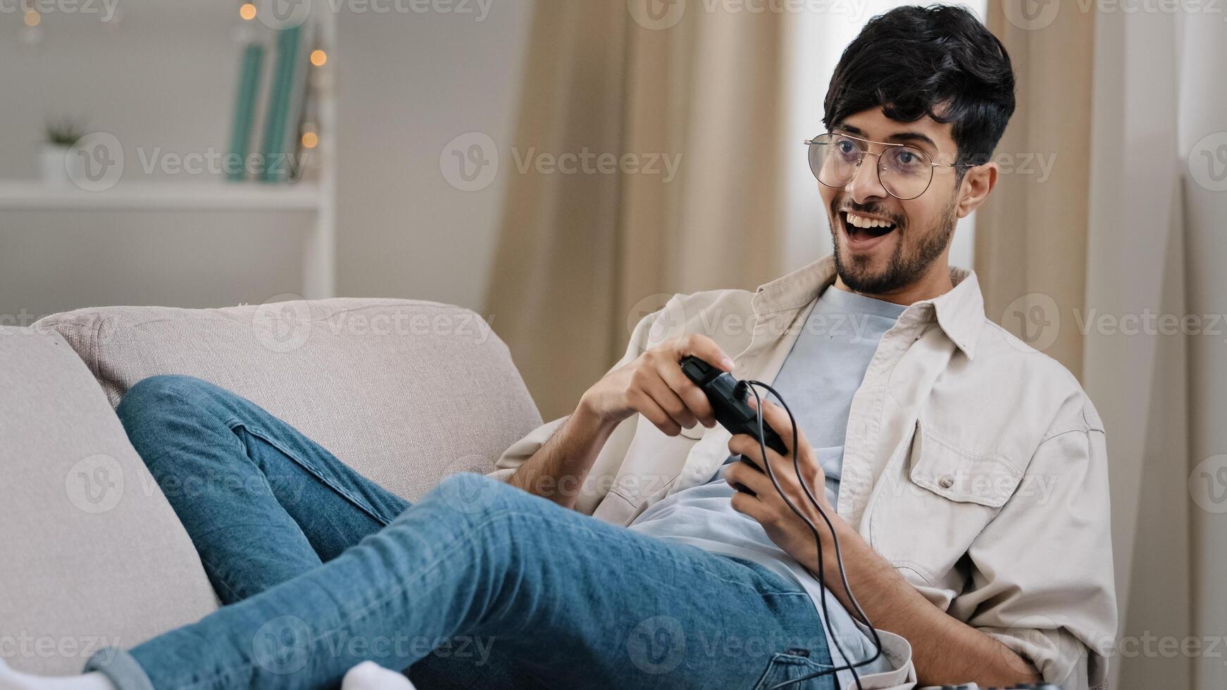 Arab guy bearded man with glasses lying on couch at home playing console online game competition emotionally winning yes hand gesture victory having fun with controller addiction to videogaming photo