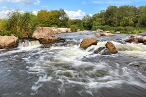 Tormentosos arroyos de agua bañan las orillas rocosas del río, superando los rápidos en un brillante día de verano. foto