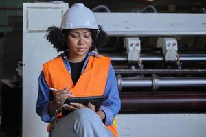 African American female engineer worker in safety uniform and hard hat, quality inspect by tablet, maintenance, check the machine at paper production manufacture industry, factory technician manager. photo