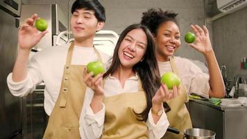 tre unga glada studenter i matlagningskurser bär förkläden njuta och glada, roliga retas med äpple och apelsin i köket, ler och skrattar, förbereder frukter för att lära sig roliga kulinariska kurser tillsammans. video