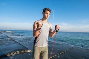 Young fit male training with athletic rubber on the beach in summer day photo