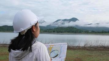 donna ecologista ingegneria in un casco con in mano una mappa si trova sulla riva di un fiume per sviluppare una diga idroelettrica per generare elettricità. concetti di energia pulita e tecnologia. video