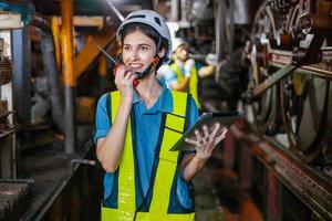 hermosa mujer ingeniero en fábrica hablando en el radio vistiendo blanco casco y trabajo ropa y mano participación tableta a lugar de trabajo foto