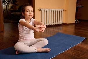 caucásico encantador niño niña 5 5 años viejo, sentado descalzo en yoga estera, extensión brazos, mirando con confianza a cámara foto