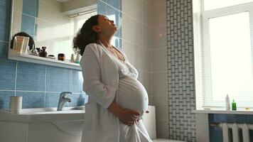 africain américain adulte Enceinte femme dans blanc peignoir de bain, permanent dans le Accueil salle de bains, souriant à la recherche de côté et doucement caressant sa ventre, profiter content des moments de sa grossesse et maternité video