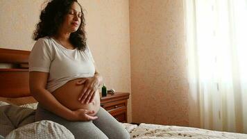 Multi etnico contento bellissima incinta donna aspettandosi un' bambino, seduta su il letto a casa Camera da letto, delicatamente carezzevole sua gonfiarsi, sperimentare contento emozioni di sua gravidanza e maternità stile di vita. video