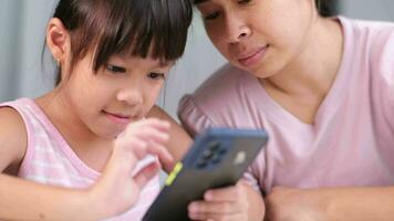 Happy Asian family mother and cute daughter using phone together at home. Mother and little girl laughing happily using smartphone watching funny social media video app sitting at table together.