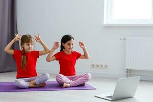 dos niñas practicando yoga, estiramientos, fitness por video en el cuaderno. entrenamiento de educación en línea distante, aeróbico en casa. estilo de vida saludable, coronavirus, quédate en casa. los niños hacen deporte en casa en cuarentena. foto