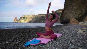 Jeune femme avec noir cheveux, aptitude instructeur dans leggings et hauts Faire élongation et pilates sur yoga tapis près le mer. femelle aptitude yoga routine concept en bonne santé mode de vie harmonie et méditation video