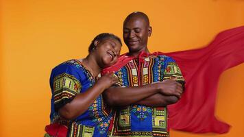 africano americano mujer presentación su marido como un héroe, vistiendo rojo capa y posando con doblada brazos. confidente hombre en cómic libro superhéroe capa siendo orgulloso de acuerdo su compañero contento. video