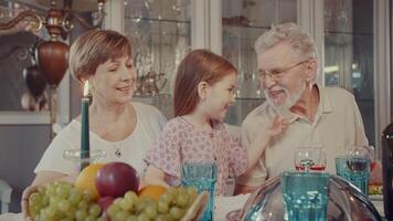 At a family dinner, a little granddaughter plays with her grandfather, pulling his beard. A girl and an elderly man are joking at the family table. Good mood. the story of one happy family. video