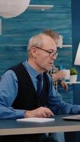 vertical . multitarefa aposentado homem lendo em computador portátil e escrevendo em caderno trabalhando a partir de lar. focado velho controlo remoto empregado procurando, analisando financeiro relatórios enquanto idosos esposa assistindo televisão dentro fundo video