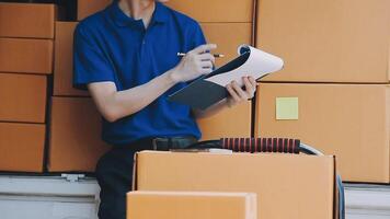 Young cheerful delivery man with parcel near car full of cardboard boxes, full length shot. Shipping service concept video