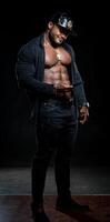 fuerte atleta en oscuro pantalones poses a el cámara en negro antecedentes. desabrochado negro camisa y Perfecto abdominales. retrato de un afro americano carrocero con teléfono en manos. foto