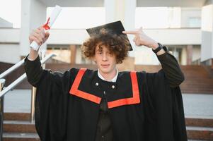 Low angle portrait of happy triumphant male graduate standing near university holding up diploma. From below of young handsome man proud of academic achievements celebrating college graduation photo