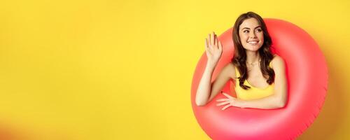 Happy young woman wearing pink swimming ring, waving hand and saying hi, enjoying vacation, summer beach holiday, standing over yellow background photo