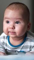 Beautiful little toddler lies on the bed on his belly. Sweet kid grimacing funnily and looking up. Close up. Blurred backdrop. Vertical video