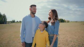 Happy adorable Caucasian loving family couple male man father woman mother female parents hugging posing together hug cuddle adopted little boy child kid son looking camera outside nature park field video