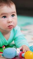charmant bébé garçon dans bleu barboteuse mensonges sur coloré tapis. mignonne enfant en jouant avec une jouet, tirant il à le sien bouche et à la recherche à il avec intérêt. verticale video