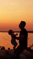 voorzichtig vader spelen met zijn baby Bij de rivier- waterkant. liefhebbend vader gooien omhoog zijn zoon en kind verheugt zich. instelling zon over- water Bij achtergrond. verticaal video