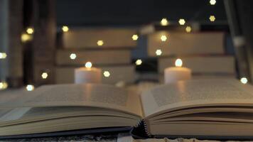 Open book changing focus with burning candles and fairy lights in the background. A pile of books is blurred in the background video