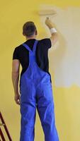 Painter doing the makeover indoors. Male worker painting wall with a paint roller next to the ladder. Improvement of a room interior. Back view. Vertical video