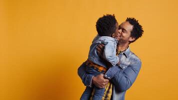African american dad caressing and kissing his adorable baby boy in studio, carrying his toddler and playing around with him. Cute family showing love and devotion to his infant son. Camera B. video