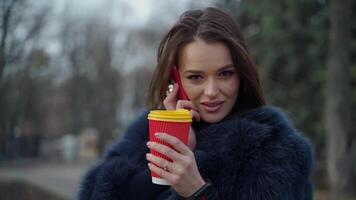 attraente ragazza all'aperto. giovane donna nel pelliccia cappotto parlando il Telefono mentre potabile caffè nel il parco. ritratto di un' femmina con plastica tazza. lento movimento. video