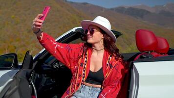 Young girl makes selfie with smartphone sits in convertible car among mountains. video