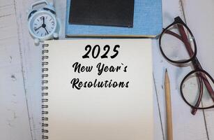 A workspace featuring a notepad with 2025 New Year's resolutions, glasses, a pen, books, and clock photo