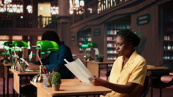 concentré africain américain étudiant est assis dans une silencieux bibliothèque en train de lire papiers et classe Remarques, en train d'étudier en dessous de le chaud lueur de une vert lampe. femme entouré par livres et apprentissage outils. caméra un. video