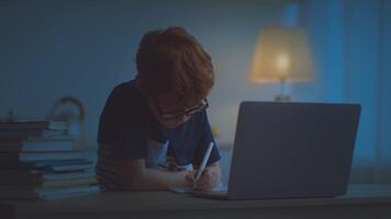 A preteen boy with ginger hair is engaged in his online studies at night. He works diligently at a desk illuminated by a warm lamp while focusing on his laptop, taking notes with a pen. video