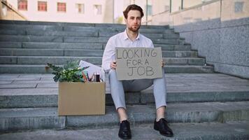 A Caucasian millennial man sits on outdoor steps, holding a sign that reads Looking for a job. Beside him, a cardboard box contains plants and office supplies, illustrating his job search. video