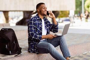 Good Business Talk. Handsome black man talking on smart phone and using laptop while working in the street photo
