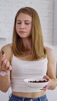 Young woman eating at home. Beautiful female standing in the modern kitchen and eating natural nuts. Healthy food concept. Vertical video