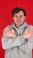 portrait worried freezing young adult curly man in gray hoodie. he rubs himself with hands to keep warm. isolated red background studio shot. concept - cold weather, heating shutdown, cold snap. video