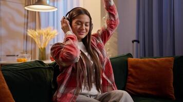 Carefree relaxed Caucasian woman teen girl happy teenager student dancing on couch sofa move dance motion listen music in wireless headphones domestic party enjoy song audio at night home apartment video