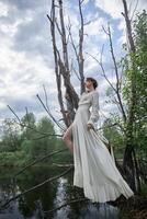 fantástico hada hermosa mujer en blanco largo vestir en antiguo seco árbol. niña princesa en pantano. Moda modelo sexy niña posando en árbol en lago. hada cuento. foto