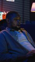 Vertical Adult man typing on laptop while he lays on a bean bag at home, engaging in freelancing and online work. Black guy multitasking efficiently and embracing the flexibility of remote job. Camera A. video