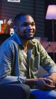 Vertical Portrait of black freelancer laying on the sofa and texting on phone, relaxing and smiling in his modern loft. Man solving daily teleworking tasks from the living room with cool lights. Camera A. video