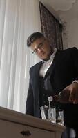 homme dans une costume verser une boisson par une ensoleillé la fenêtre. une homme portant une formel noir costume est vu verser une boisson dans des lunettes par une fenêtre avec blanc rideaux. video