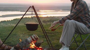 une Jeune fille, portant une plaid chemise, est cuisine nourriture dans une pot plus de une trépied tandis que randonnée dans la nature. elle est bavardage avec sa ami. portrait. video